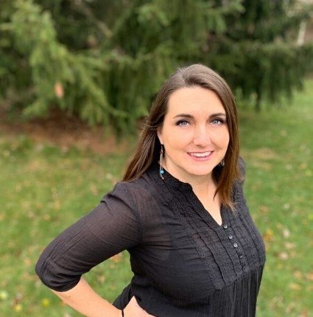 White woman in black long-sleeved blouse smiling, standing outside in front of a pine tree.