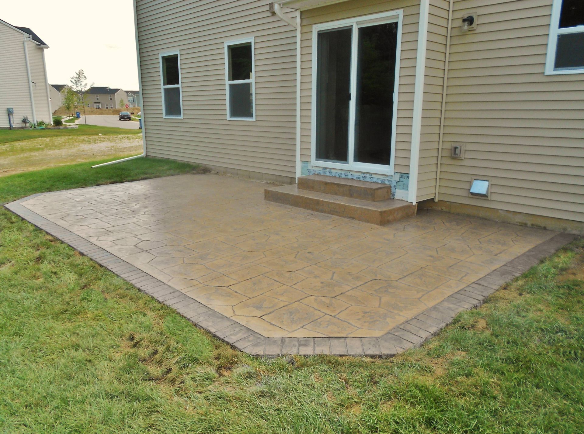 Large tan stamped-concrete patio beside a beige house with sliding glass doors and steps to the backyard