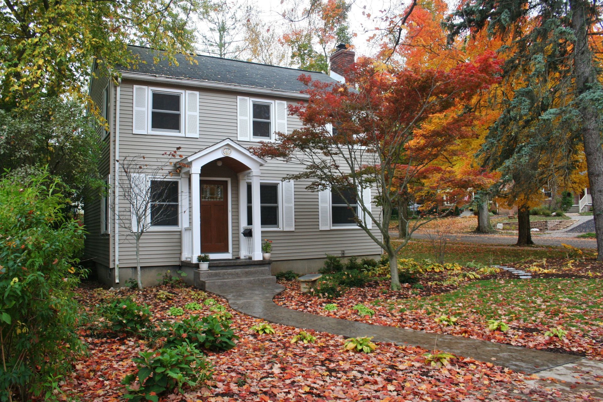 Two-story house with white siding and autumn trees, covered in fallen leaves along the walkway