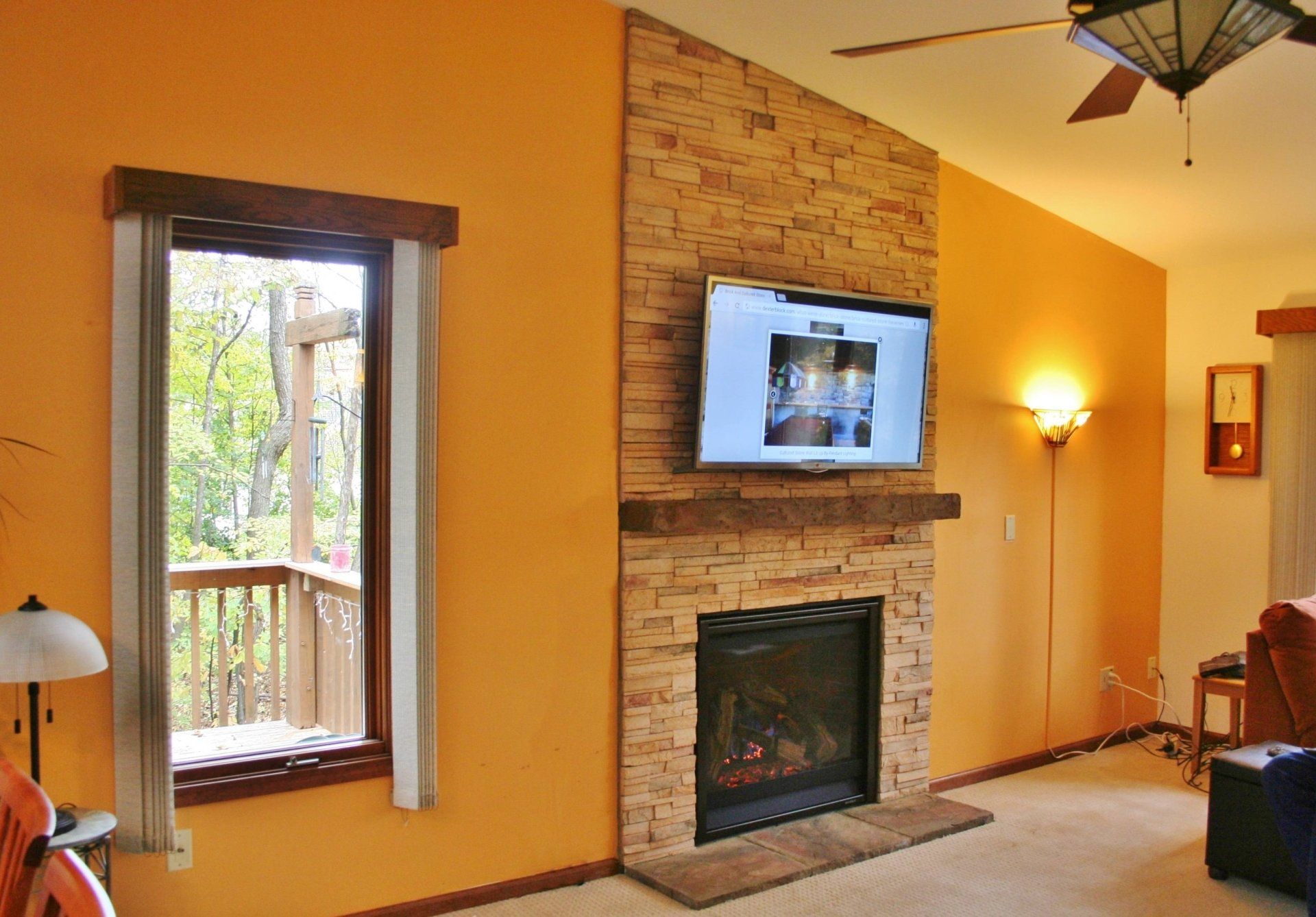 Living room with orange walls, stone fireplace, wall-mounted TV, and window with trees outside