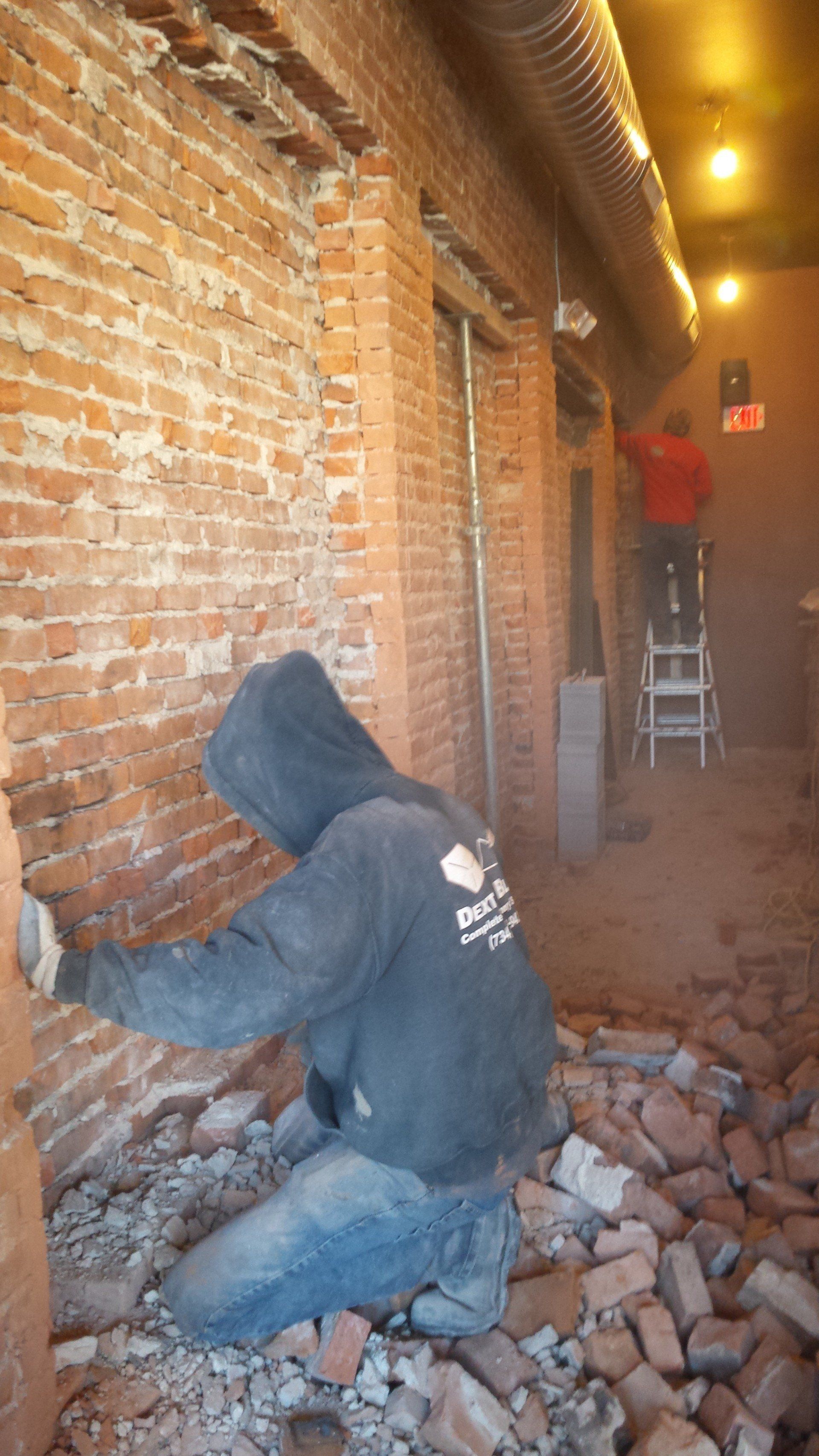 Worker in a hooded jacket kneeling on rubble, repairing a brick wall inside a dim hallway.