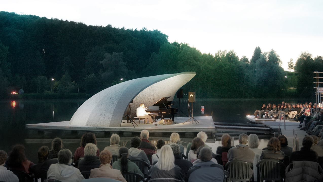 Outdoor amphitheater concert at dusk, with a white shell stage and seated audience by the water