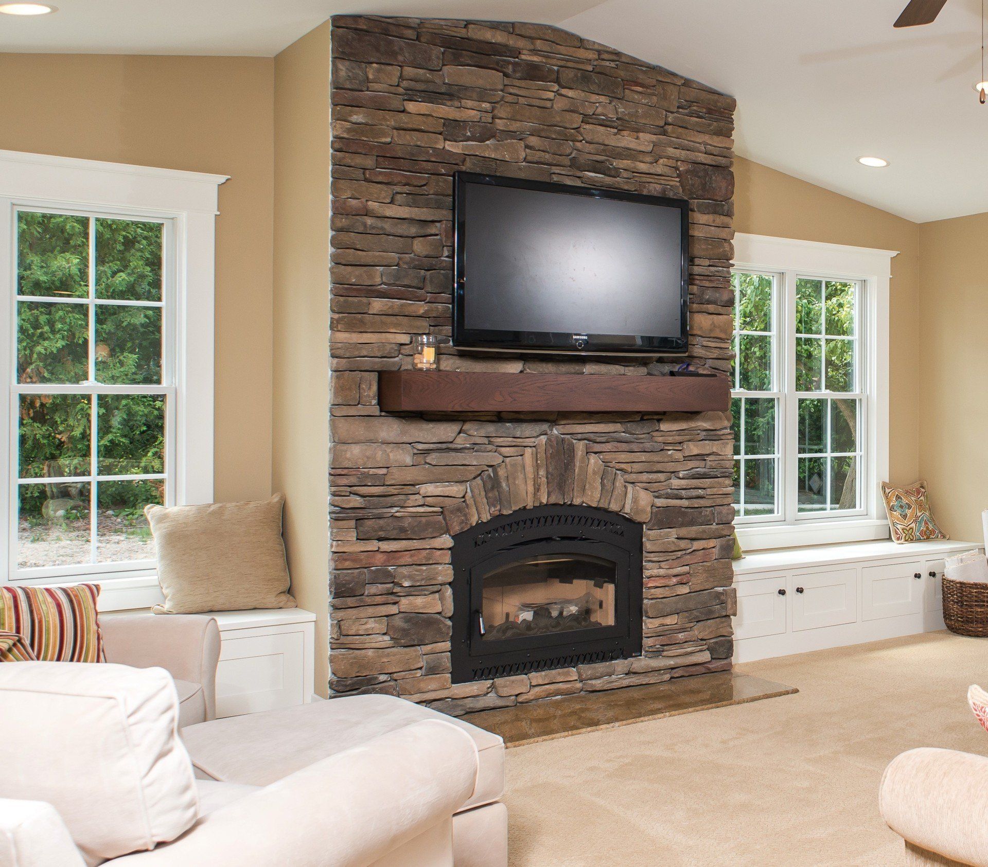 Living room with stone fireplace, mounted TV, and large windows with built-in white seating.