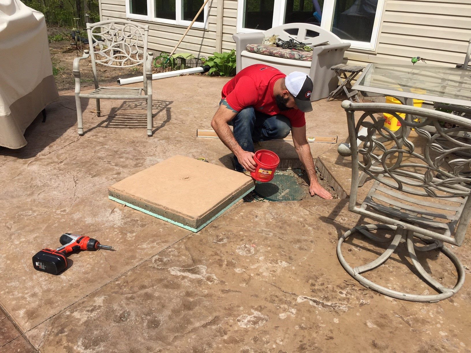 Worker kneels on patio tile, using a red tool beside stacked pavers and hoses in a backyard.