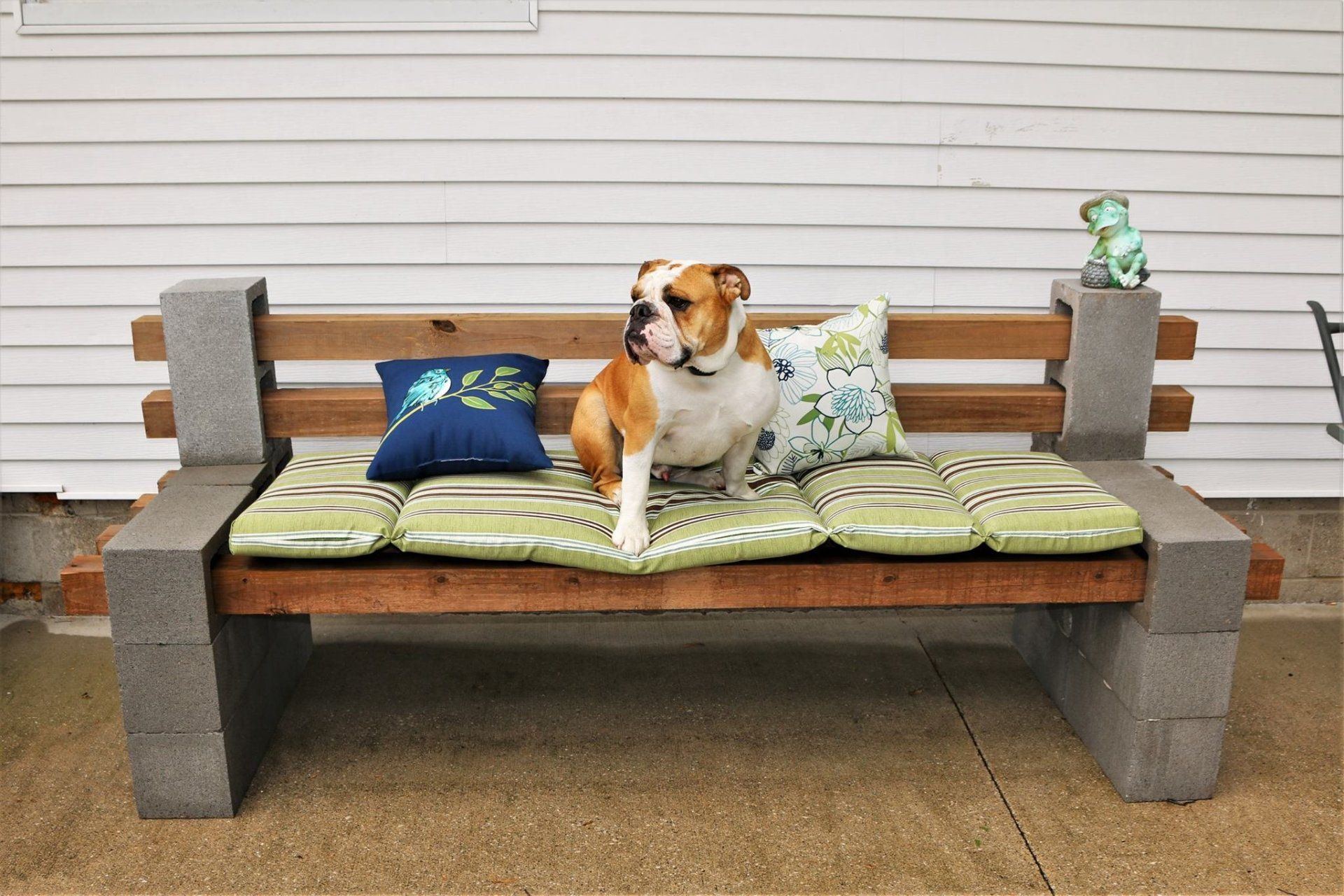 Dog sitting on a cushioned outdoor bench with patterned pillows against a white siding wall.