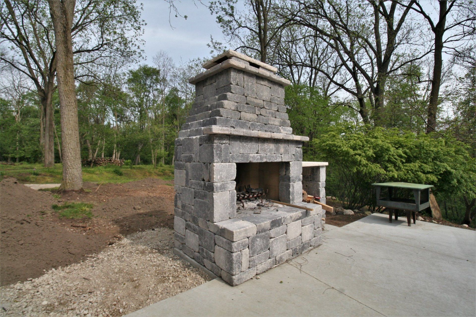 Stone outdoor fireplace beside a concrete patio in a wooded park setting
