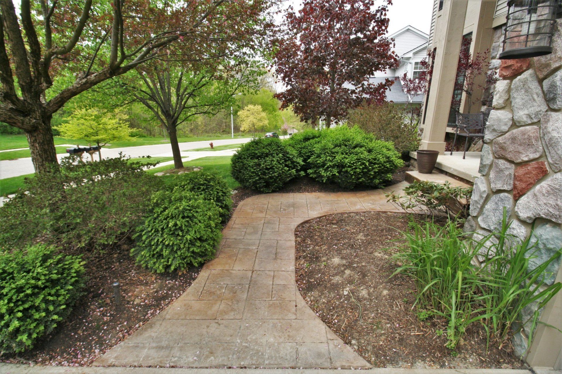 Curved concrete garden path beside shrubs, trees, and a stone house exterior.