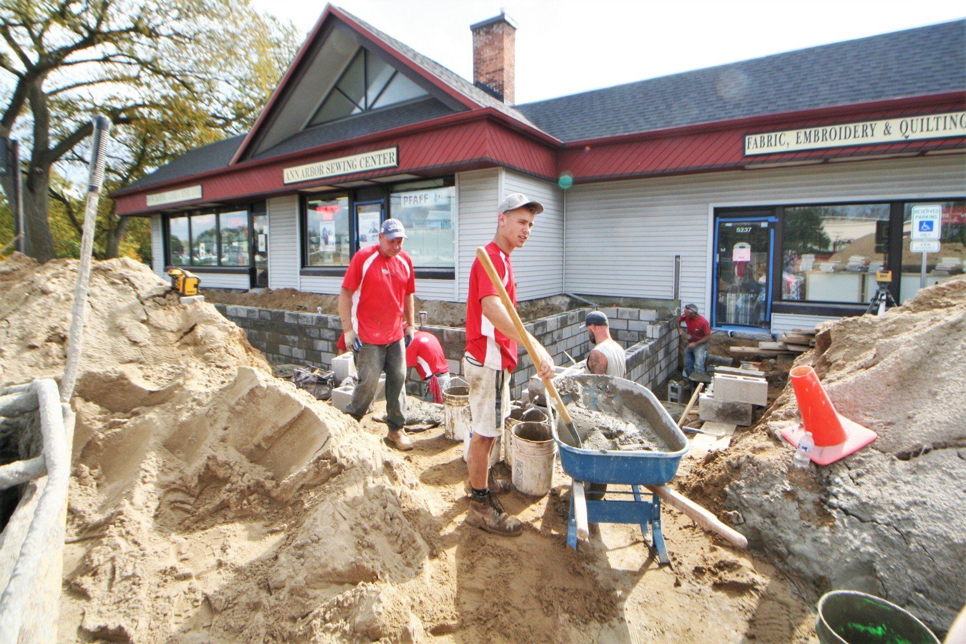 Workers filling sandbags outside a storefront beside a large sand pile and wheelbarrow