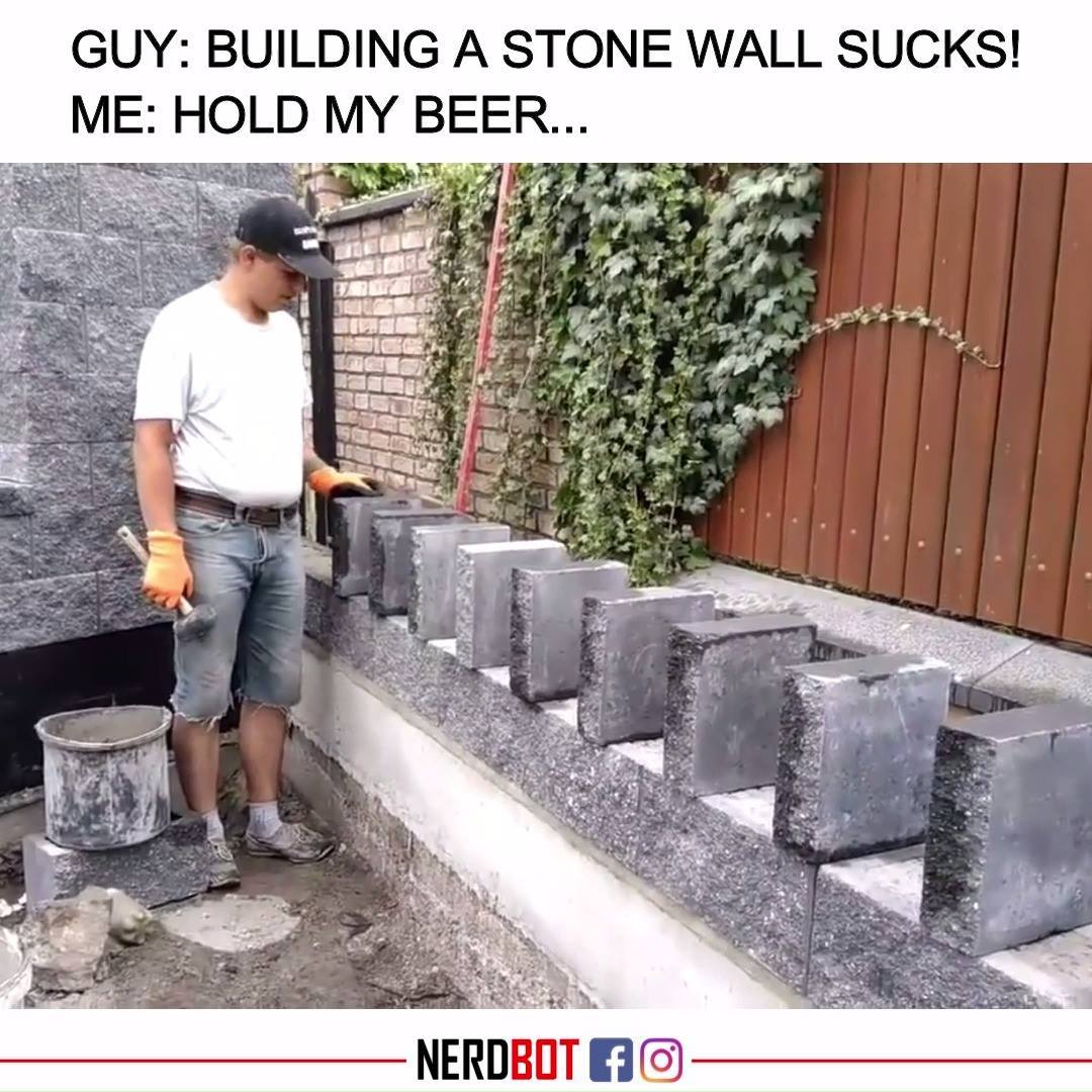 Man builds a stone wall with precise concrete blocks beside a vine-covered fence.
