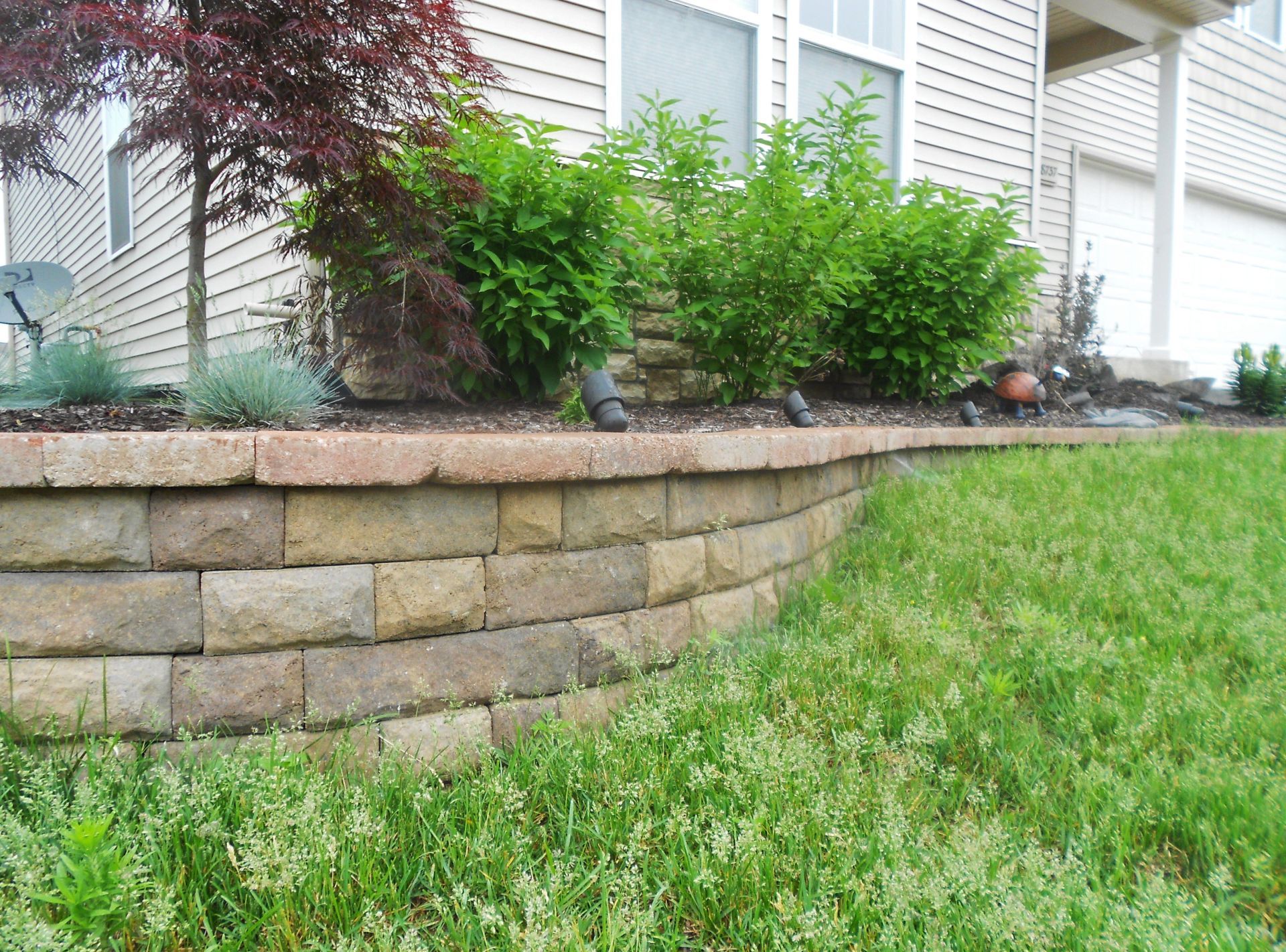 Stone retaining wall with green shrubs beside a white house and grassy lawn