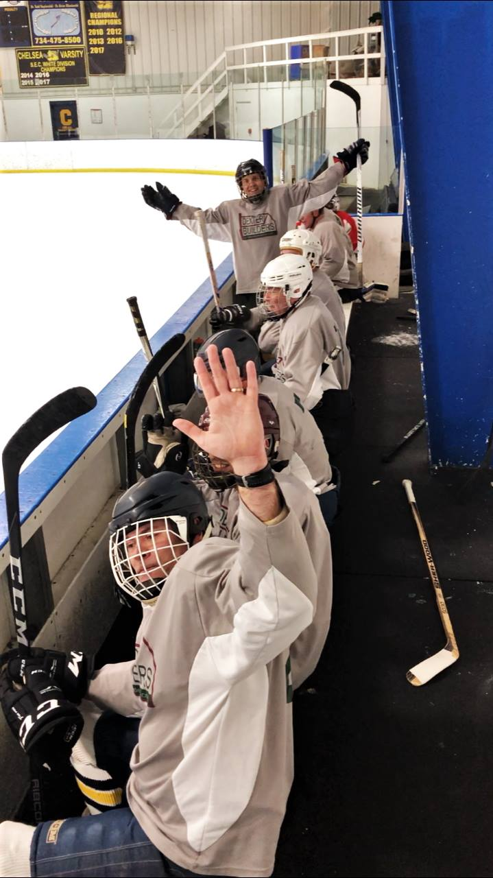 Ice hockey players in white gear lined up at the bench, one waving, inside an indoor rink