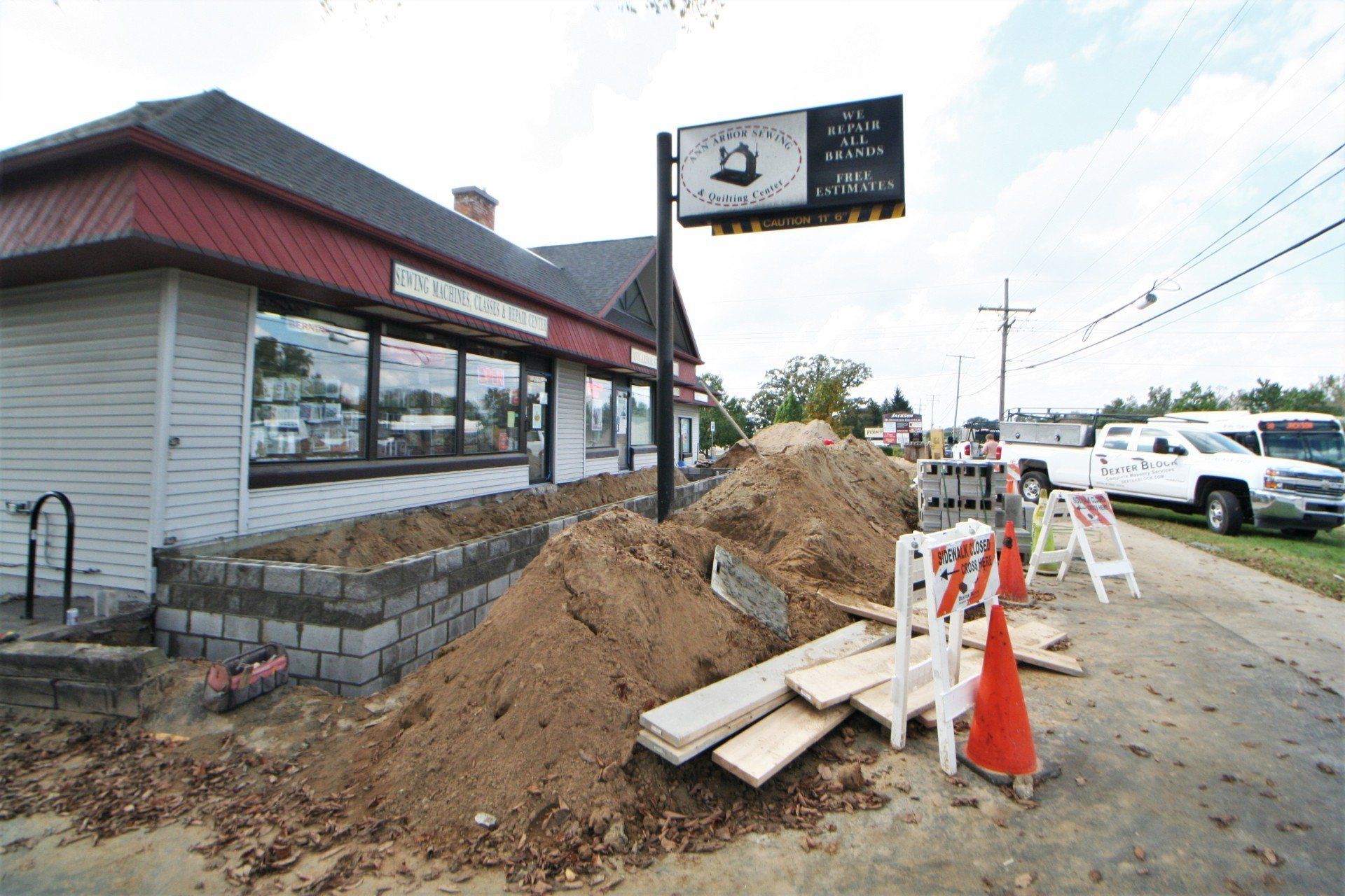 Construction trench and dirt pile beside a building and roadside, with orange cones and barricades.