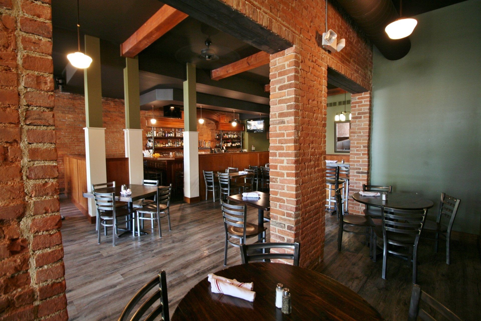 Brick-walled restaurant interior with wooden tables, black chairs, and a bar in the background