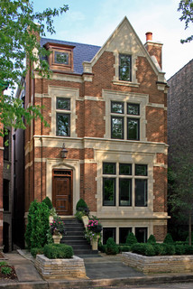 Red brick townhouse with a steep gabled roof, tall windows, and front steps along a tree-lined street