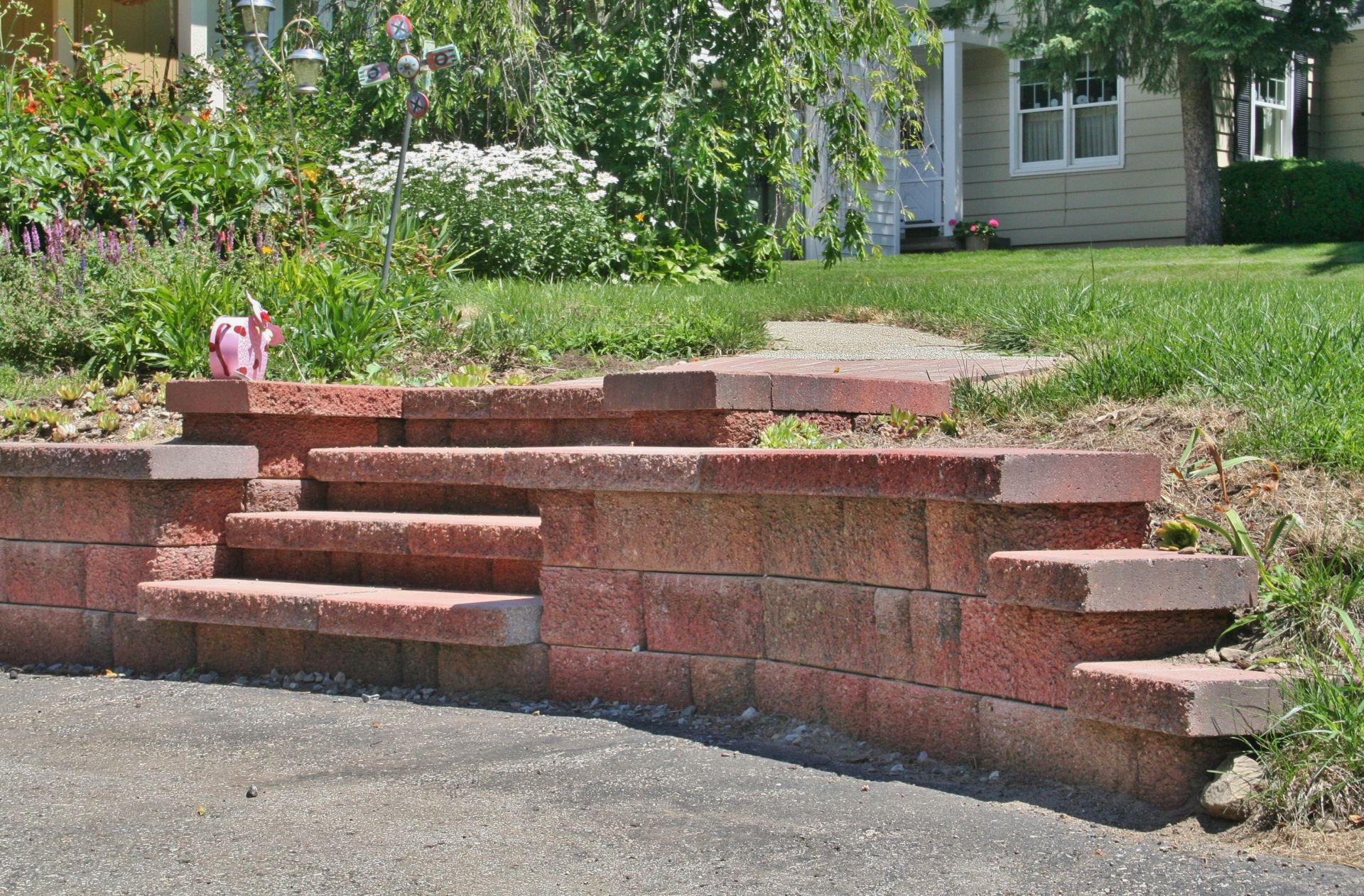 Brick retaining wall with steps beside a driveway and a grassy yard with flowers.