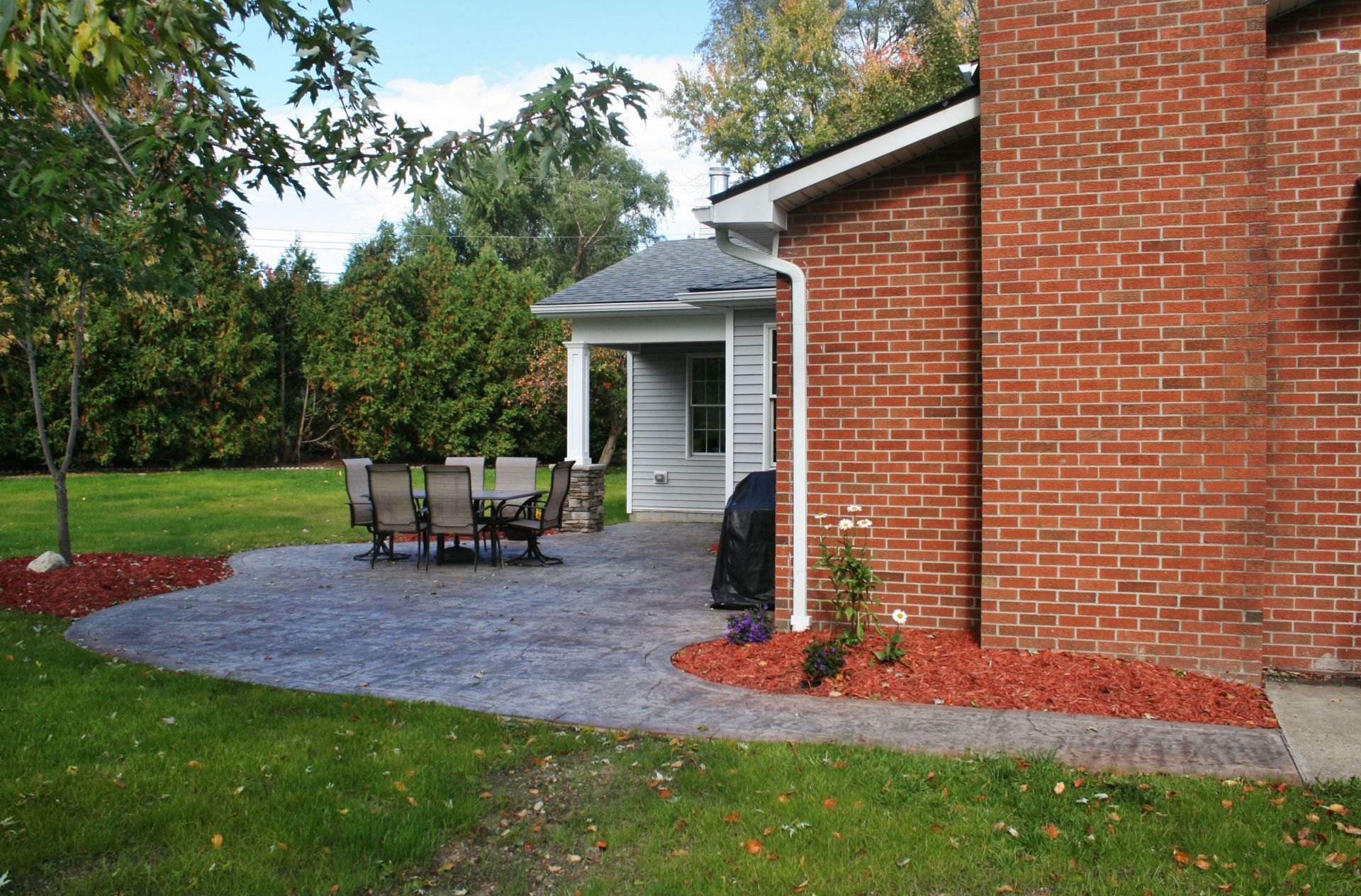 Patio with table and chairs beside a brick house, surrounded by grass, trees, and red mulch