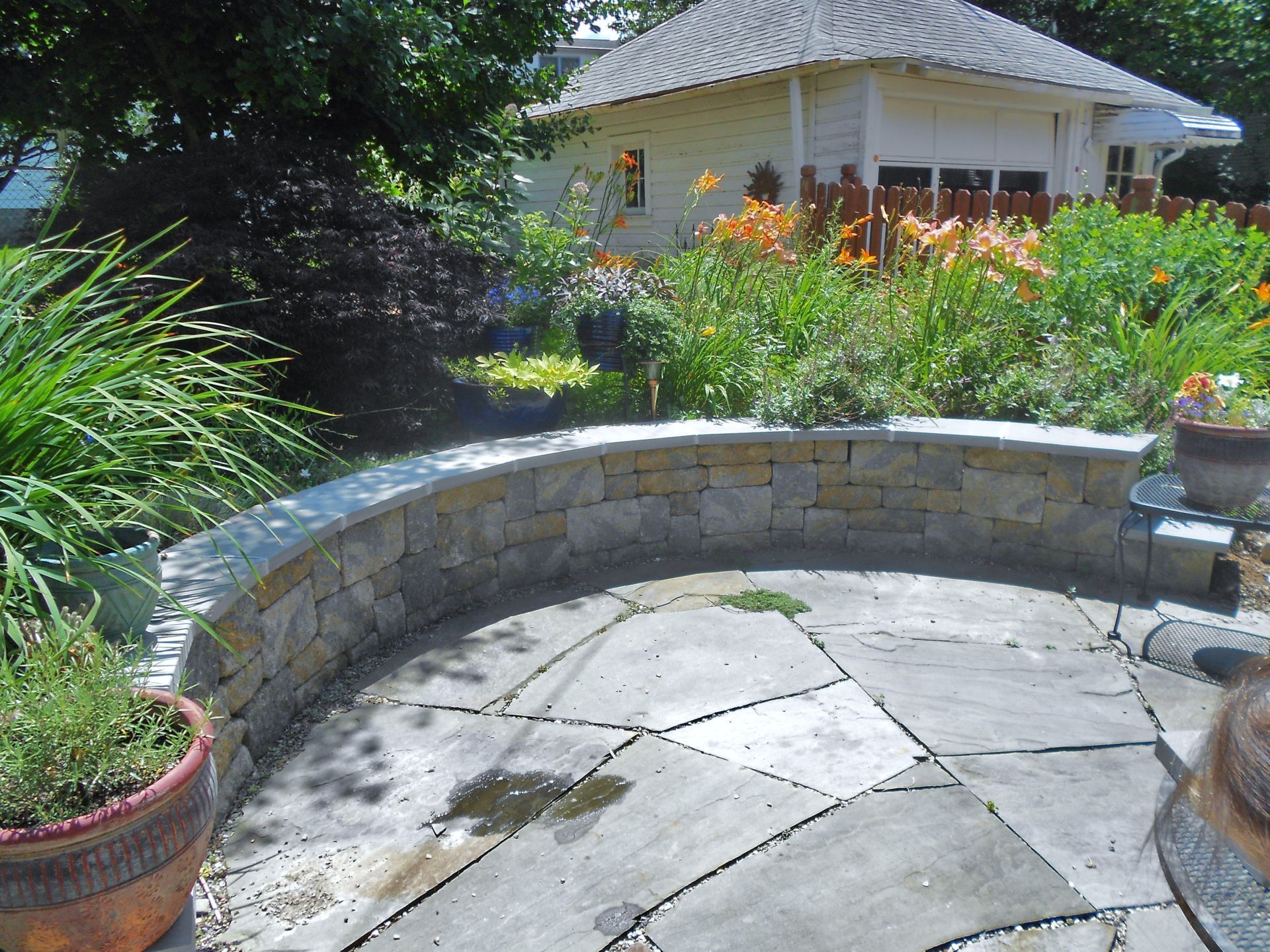 Curved stone patio beside a lush flower garden and a small white gazebo in sunlight