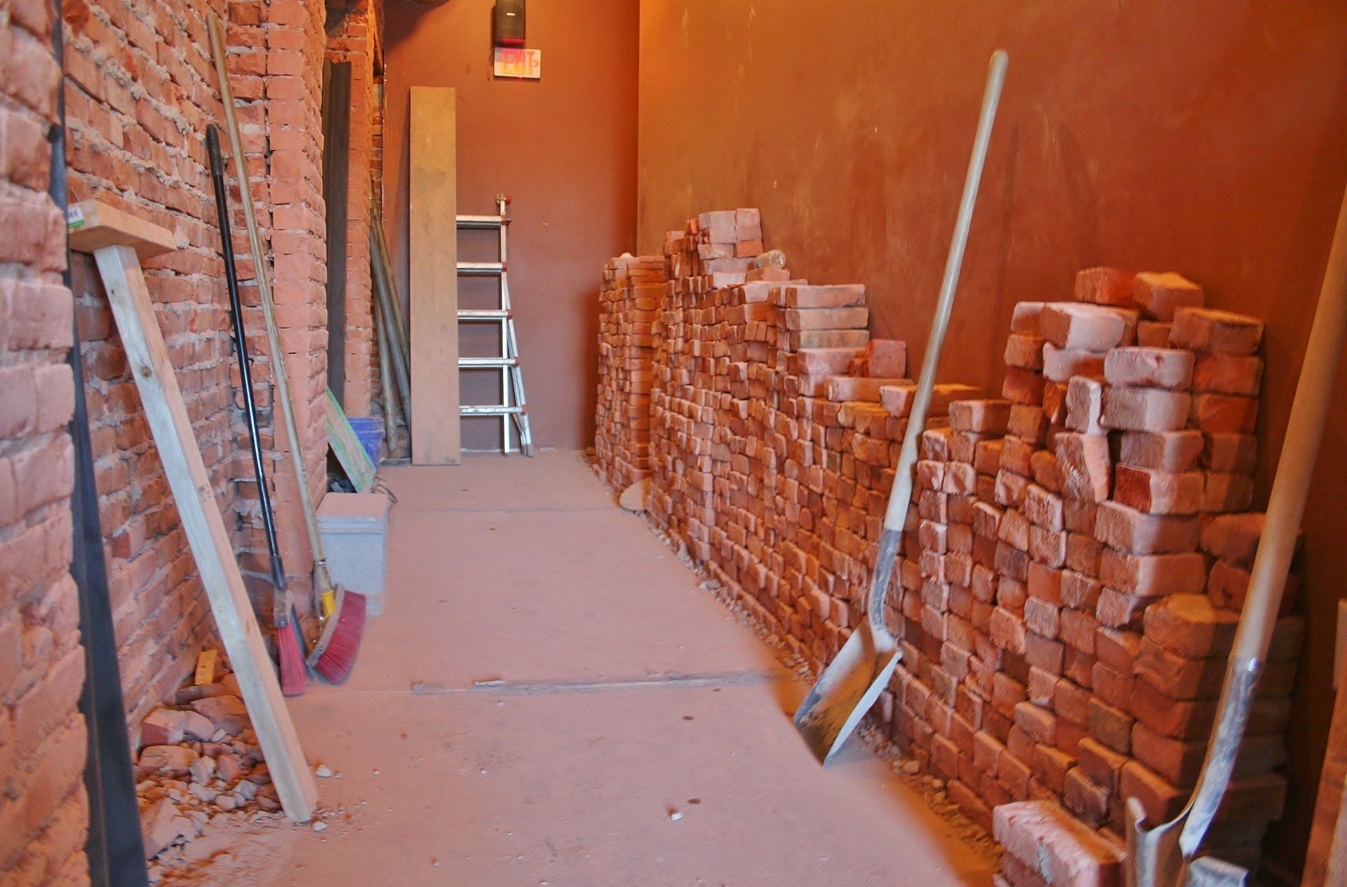Narrow construction corridor with stacked bricks, leaning ladders, and tools along unfinished brick walls.