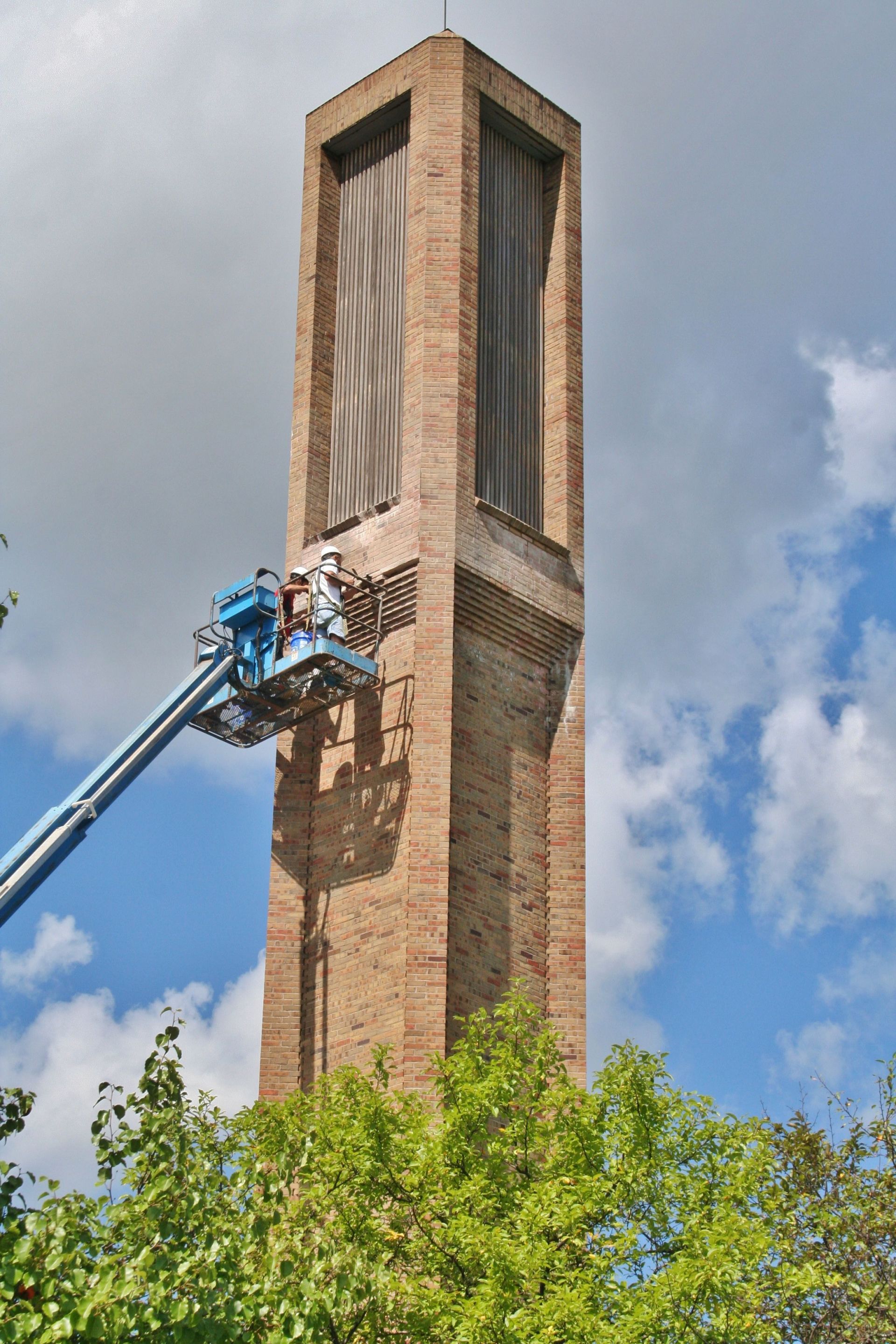 Workers on a blue lift repairing a tall brick tower against a partly cloudy sky