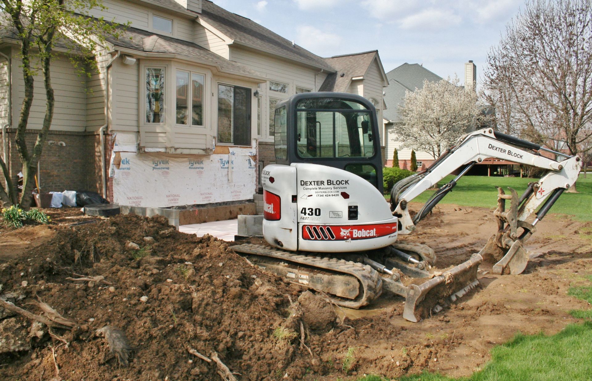 Excavator digging a trench beside a house under renovation, with exposed foundation and dirt piles.