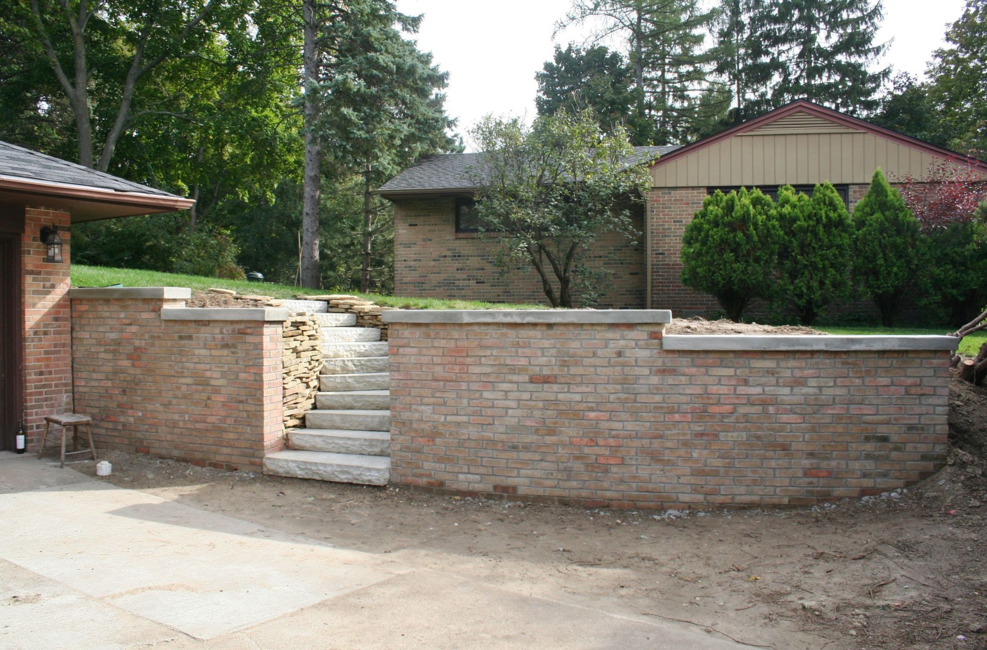 Brick retaining wall and steps leading to a raised patio in a yard with trees and a house.