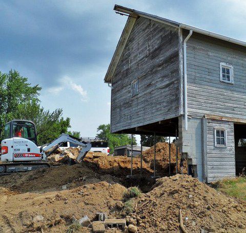 Heavy equipment excavating dirt beside a raised weathered barn under construction.