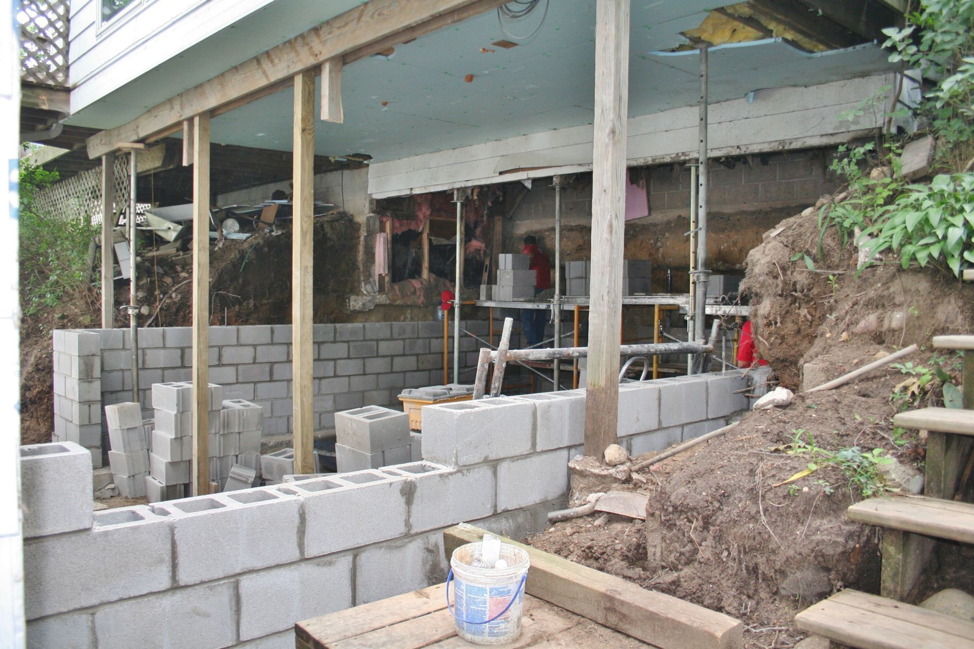 Partially built cinder block wall under a house on stilts, with construction materials and a hillside staircase