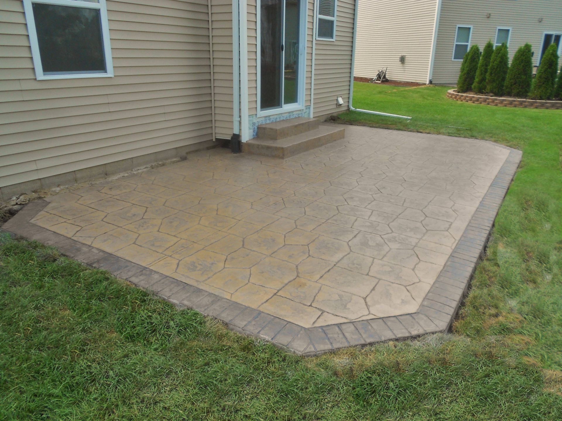 Stamped concrete patio beside a house, bordered by grass and a small step to the door.