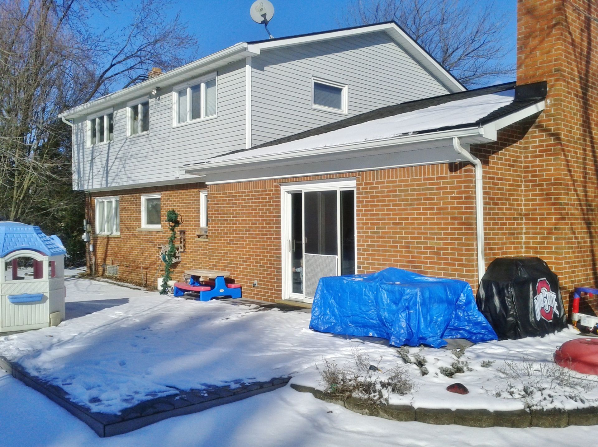 Snowy backyard beside a brick-and-gray house with blue-tarp-covered items and a grill