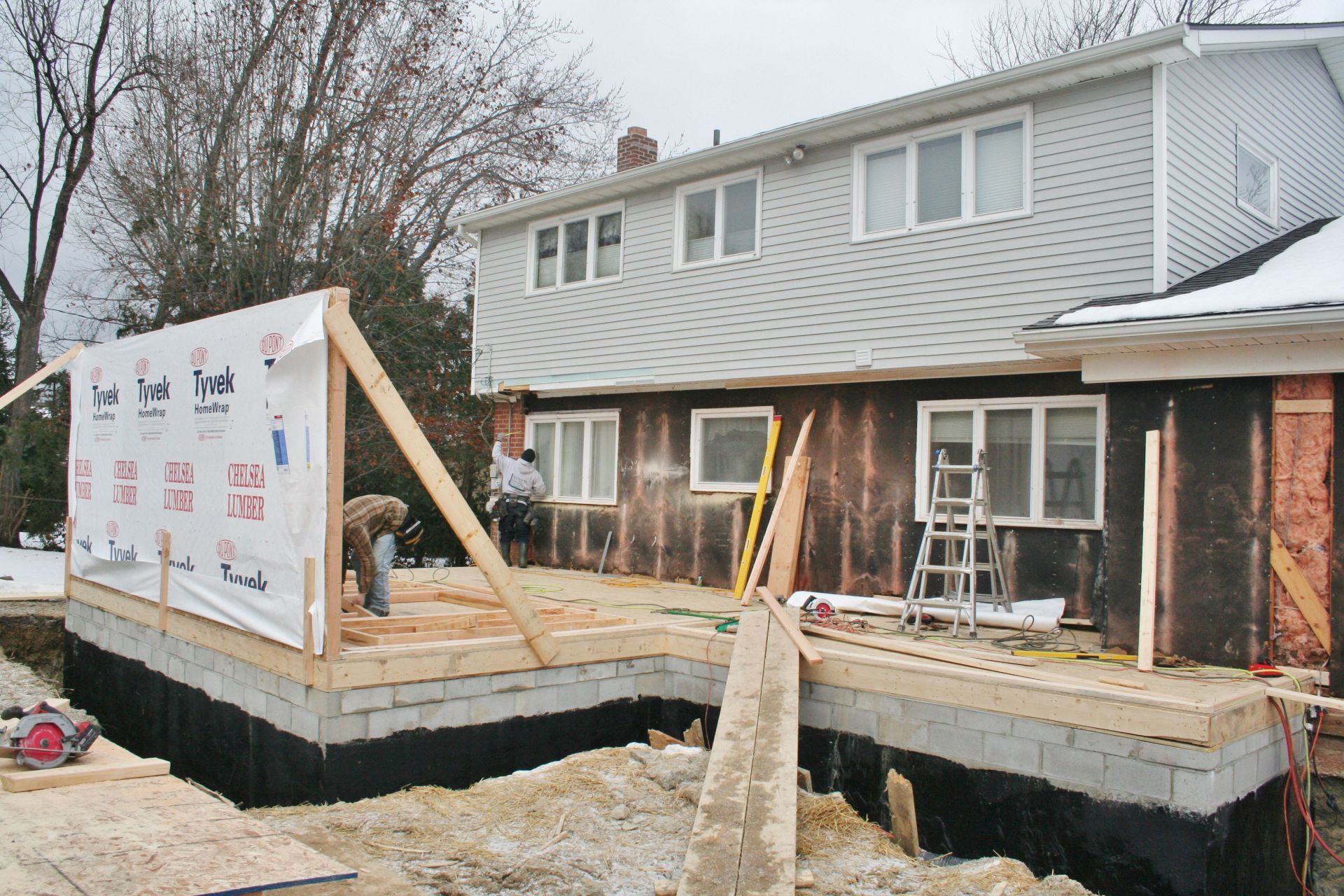 House under construction with raised wooden deck framing and workers near the foundation