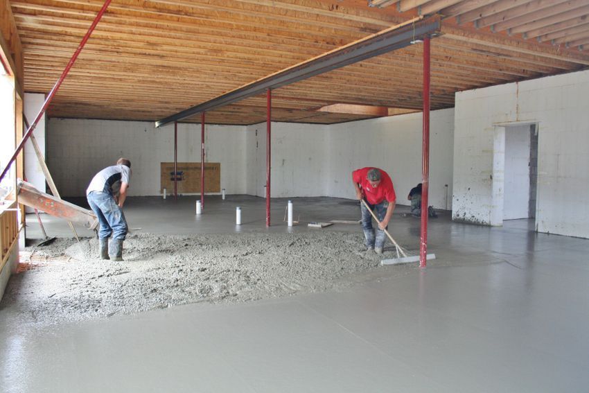 Two workers smoothing a wet concrete floor in a large unfinished room with a wooden ceiling.