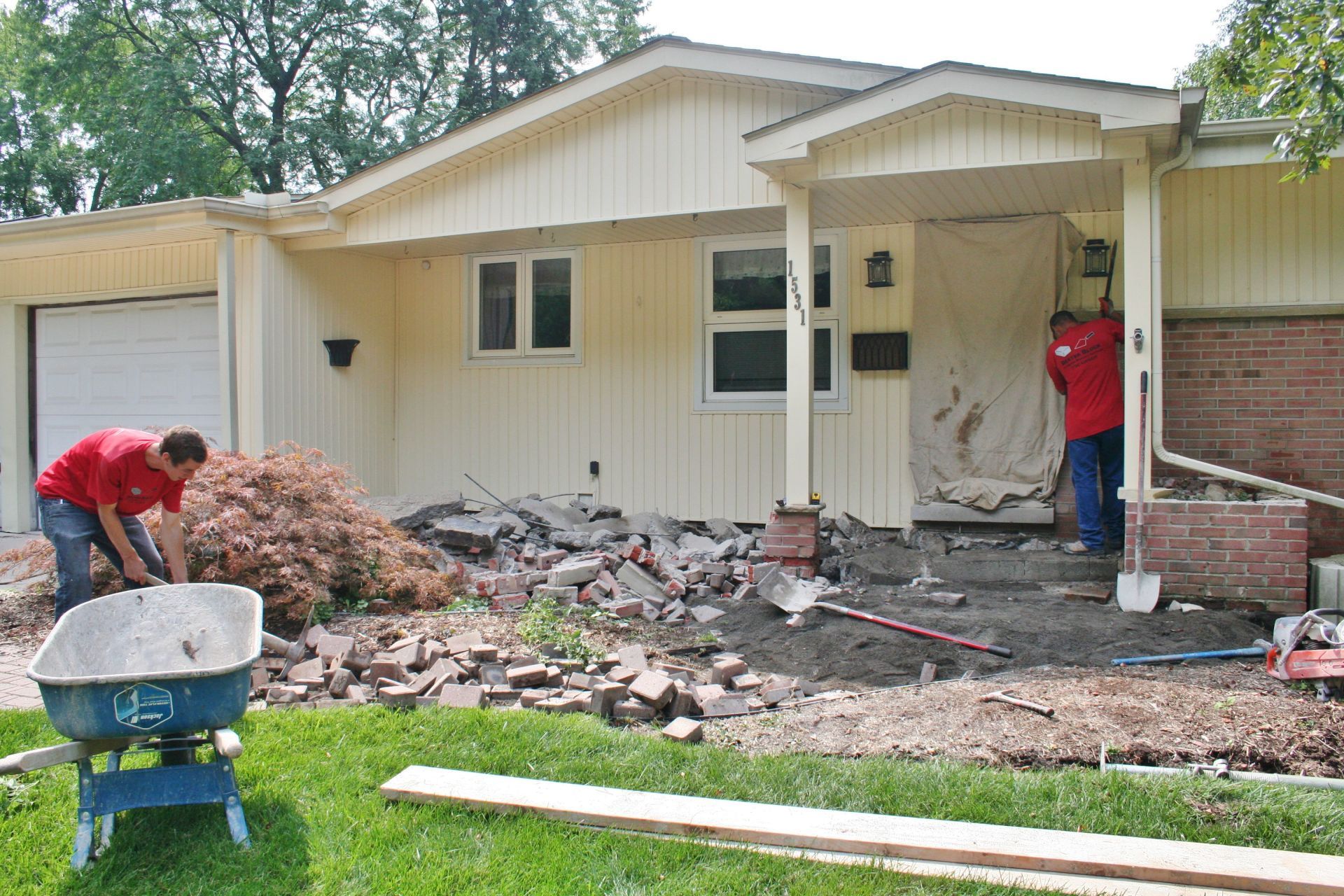 Workers removing rubble from a damaged house exterior, with debris pile and wheelbarrow in front yard