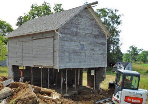 Old wooden house lifted on stilts during renovation, with a small excavator nearby.