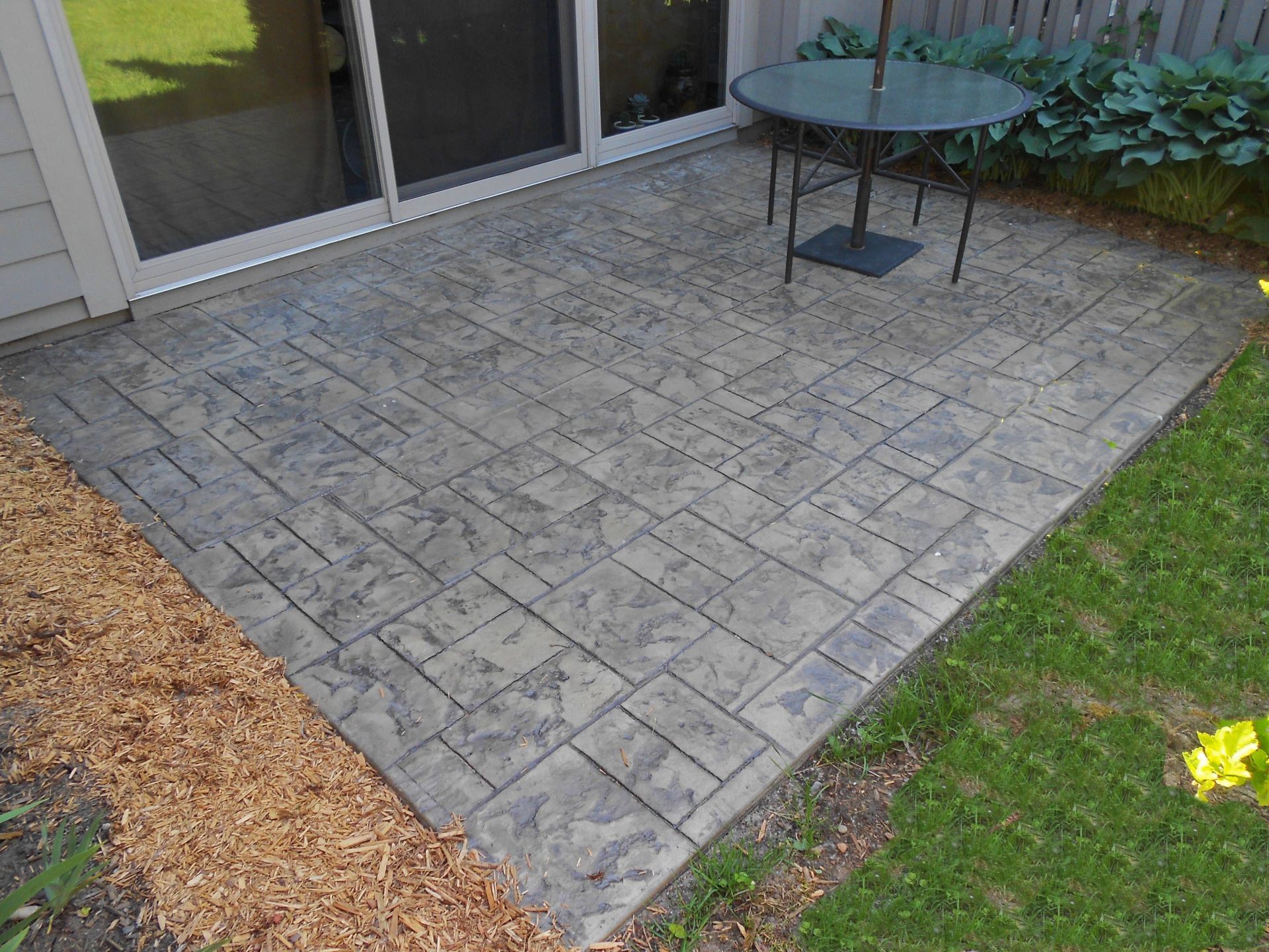 Stone patio with a round glass table beside sliding doors and a grassy yard.