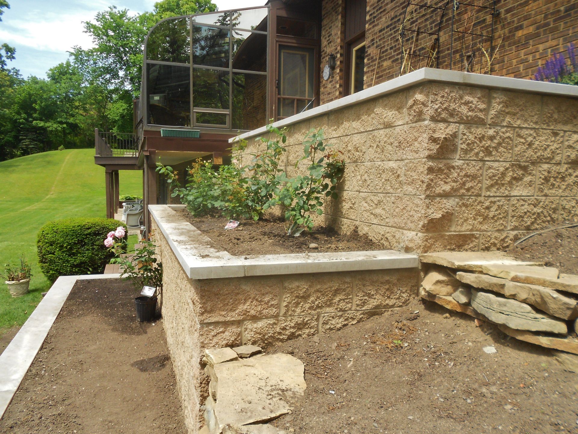 Stone retaining wall and garden bed beside a house with a balcony, bordering a sloped lawn.