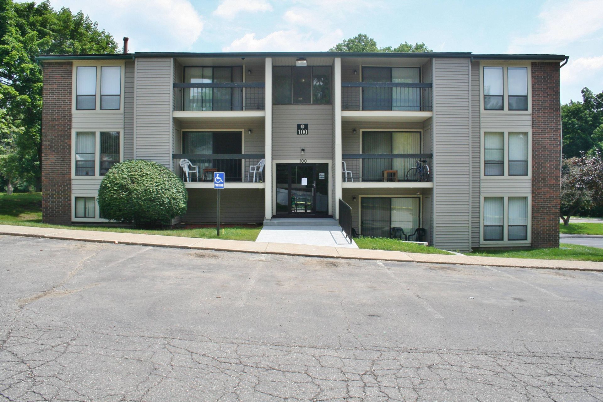 Three-story apartment building with balconies, brick sides, and a central entrance by a parking lot
