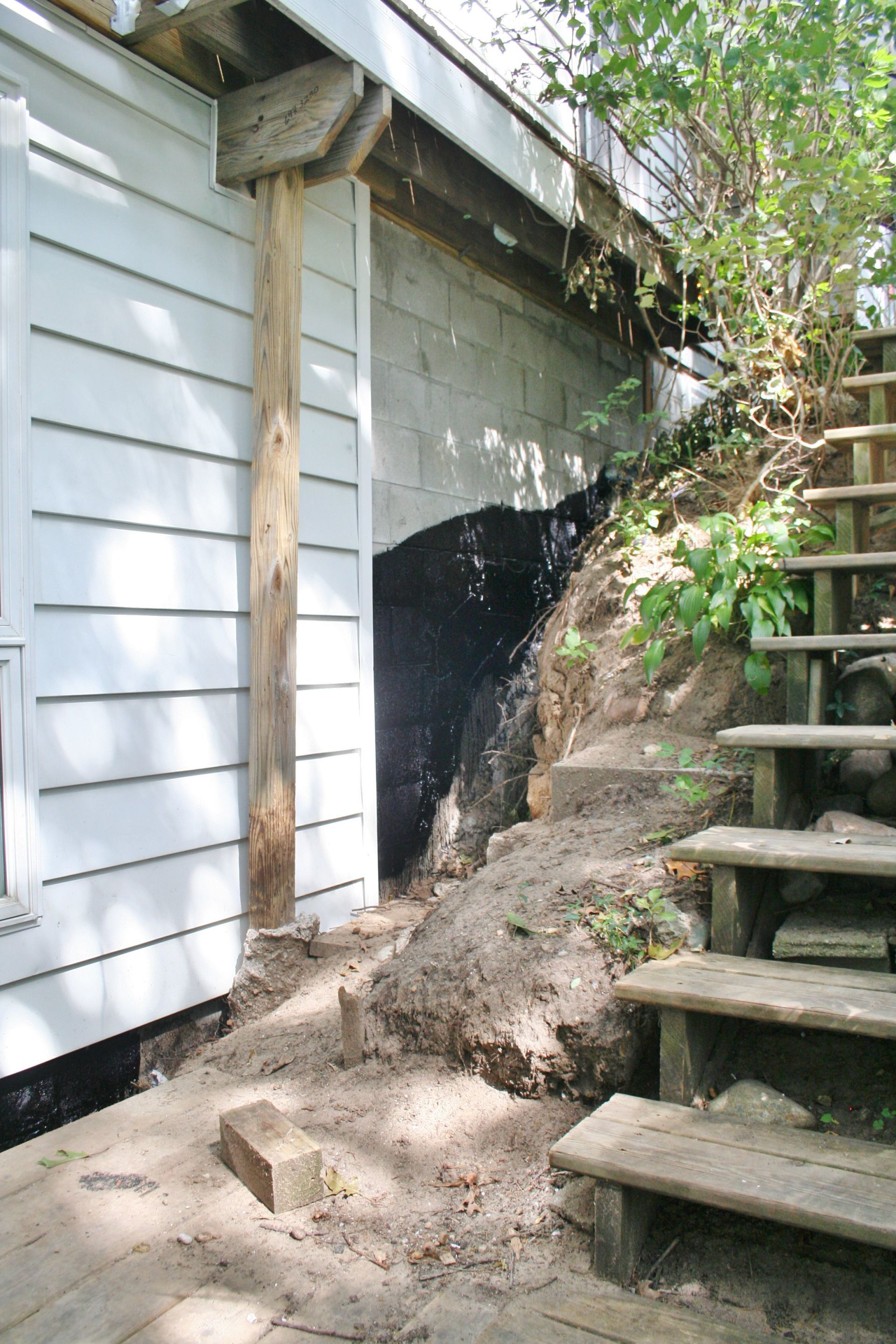 Exterior wall with exposed foundation and eroded soil beside wooden stairs and a retaining rock slope