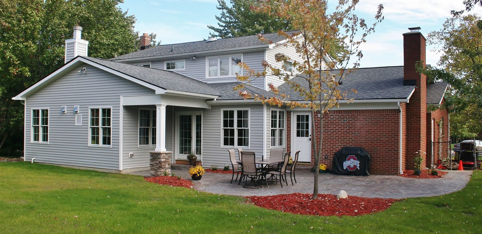 Gray single-story house with white trim, front porch, and brick chimney, set on a green lawn with autumn leaves.