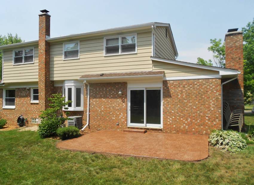 Suburban two-story brick house with tan siding, chimneys, and a brick patio in front