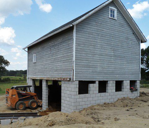 Partially built gray house on cinder blocks with a loader beside it on a dirt lot
