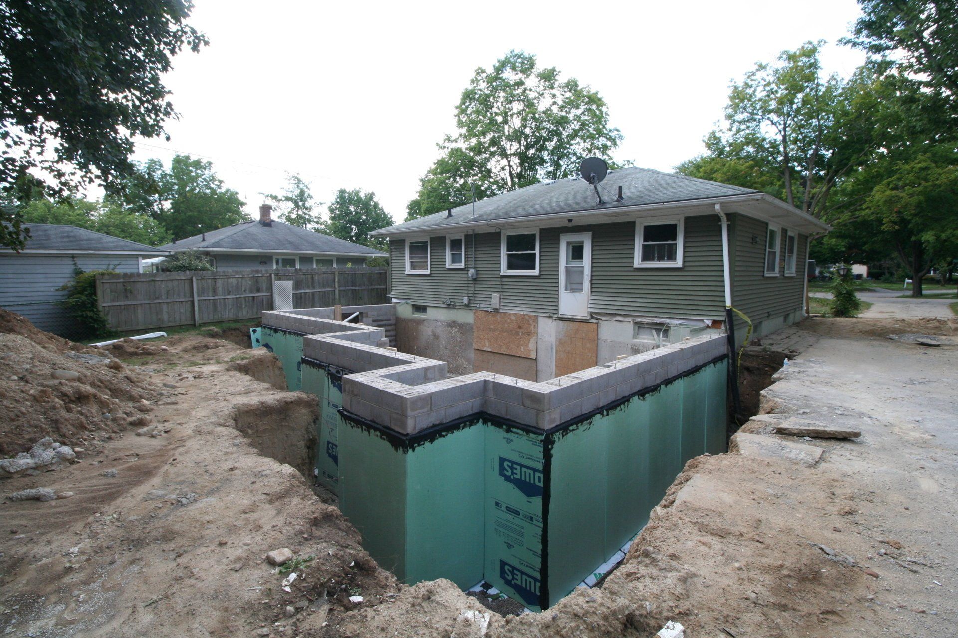 House under construction with exposed foundation and green waterproofing panels in a dirt-filled yard
