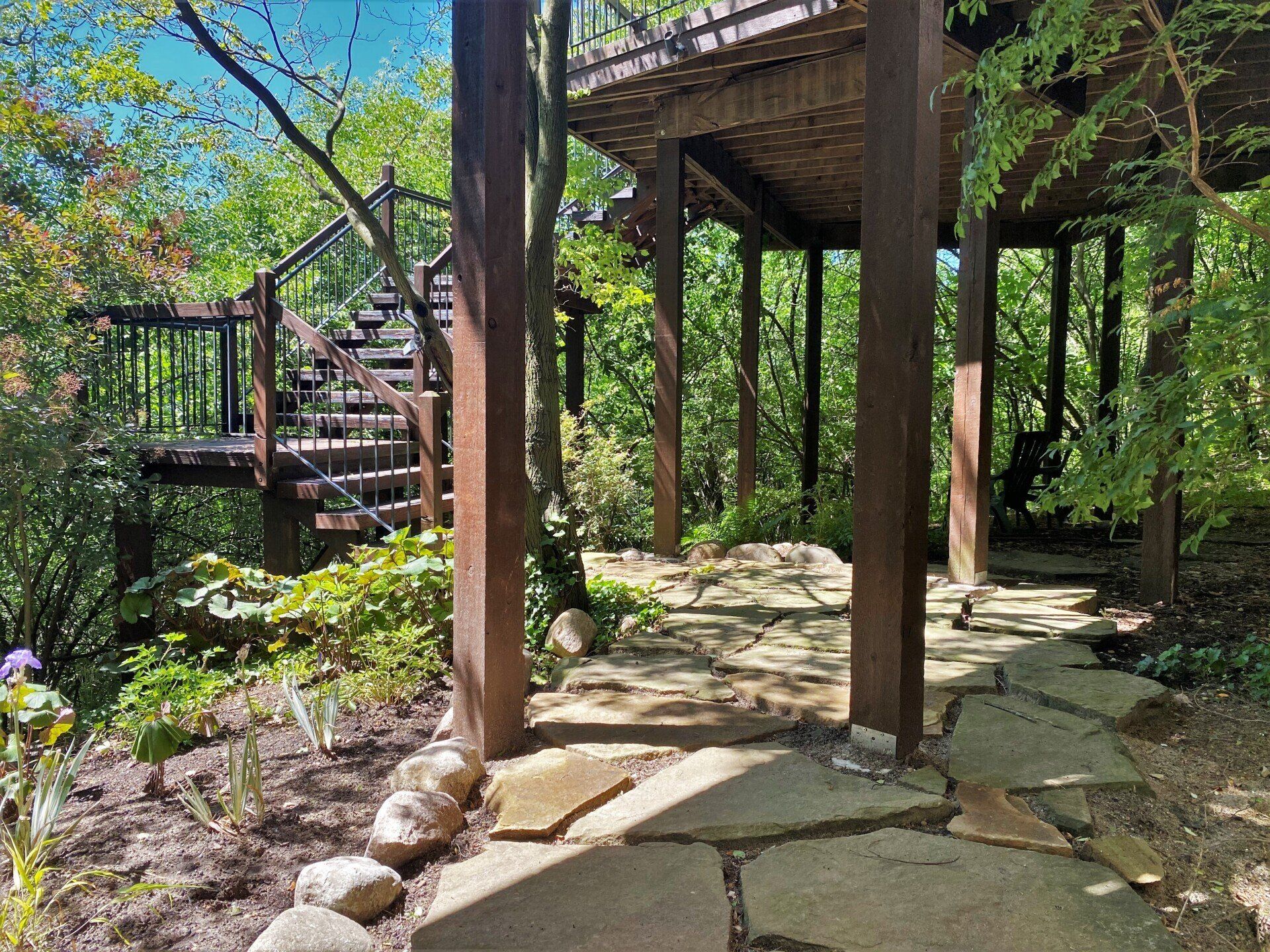 Wooden deck and stairs in a lush garden with stone path and tall posts under blue sky