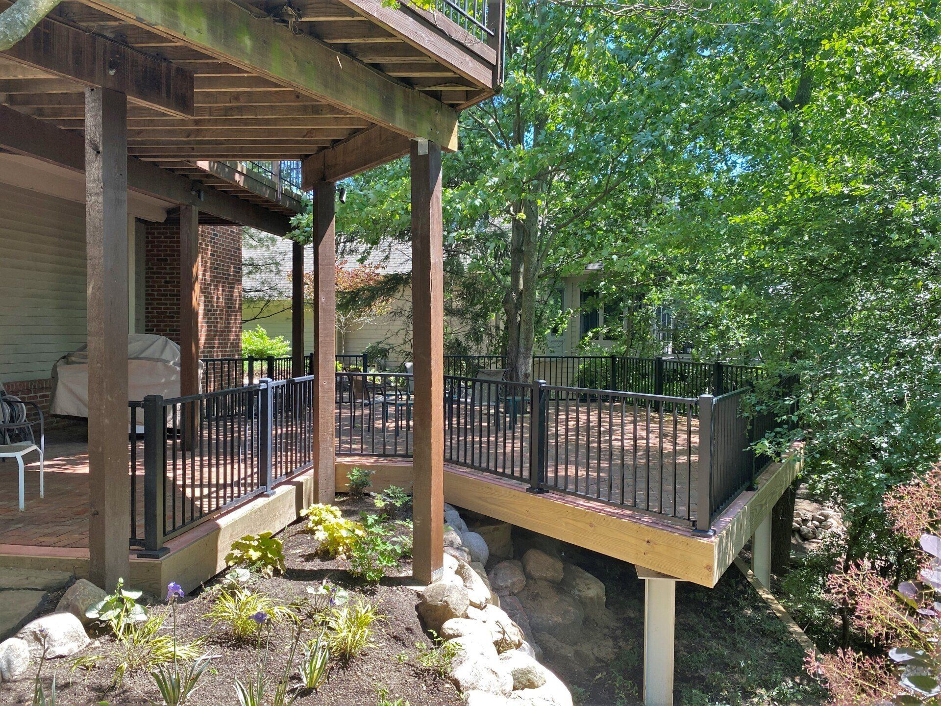 Wooden deck with black railings beside a house, overlooking a leafy hillside garden.