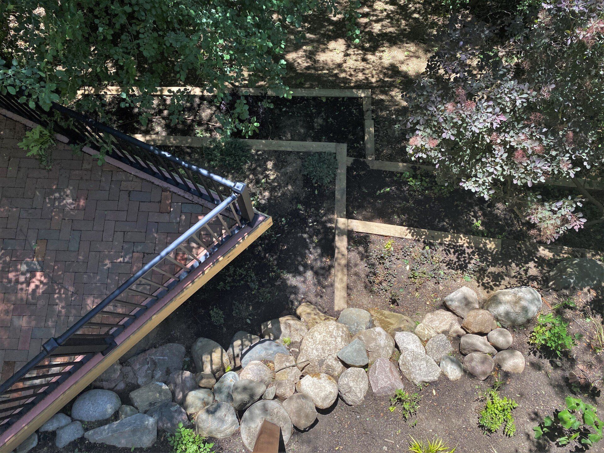 Stone-lined stream beneath a wooden bridge beside a brick path, shaded by trees.