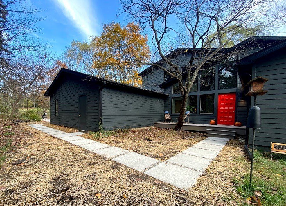 Modern house exterior with a red front door, black siding, and a stone walkway in a wooded yard