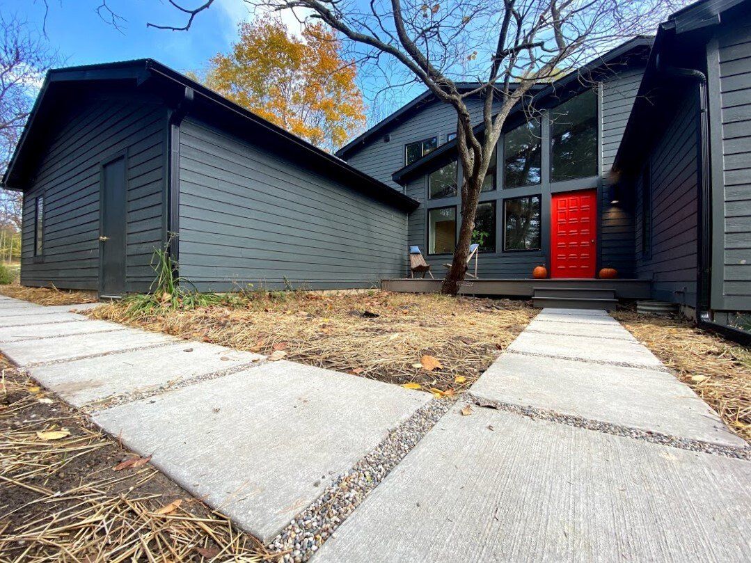 Modern house exterior with dark siding, concrete walkway, and a bright red front door under autumn trees
