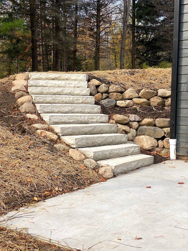 Stone steps built into a landscaped slope beside a concrete driveway, with trees in the background