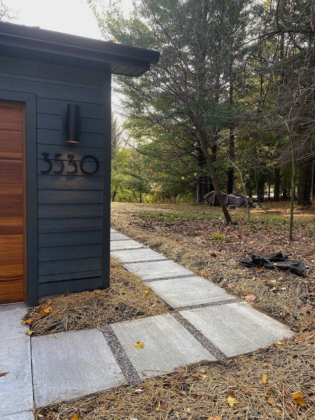 Dark gray house exterior with wood door, number 3530, and a concrete path through a wooded yard.