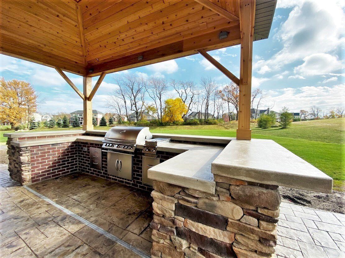 Covered outdoor stone grill area with built-in barbecue and grassy backyard view