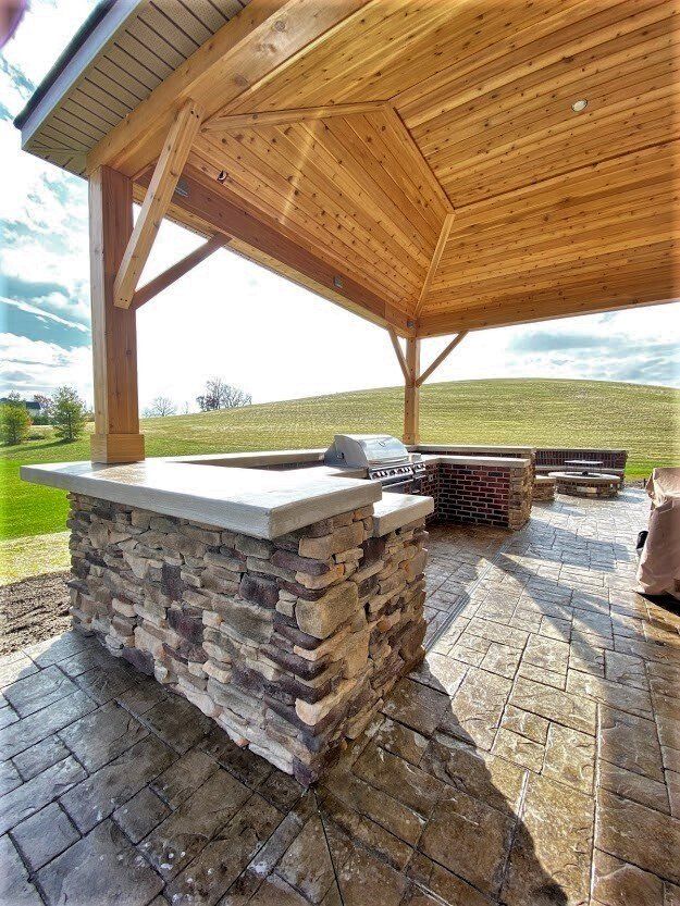 Stone outdoor pavilion with wooden roof and built-in grill on a paved patio overlooking a grassy field